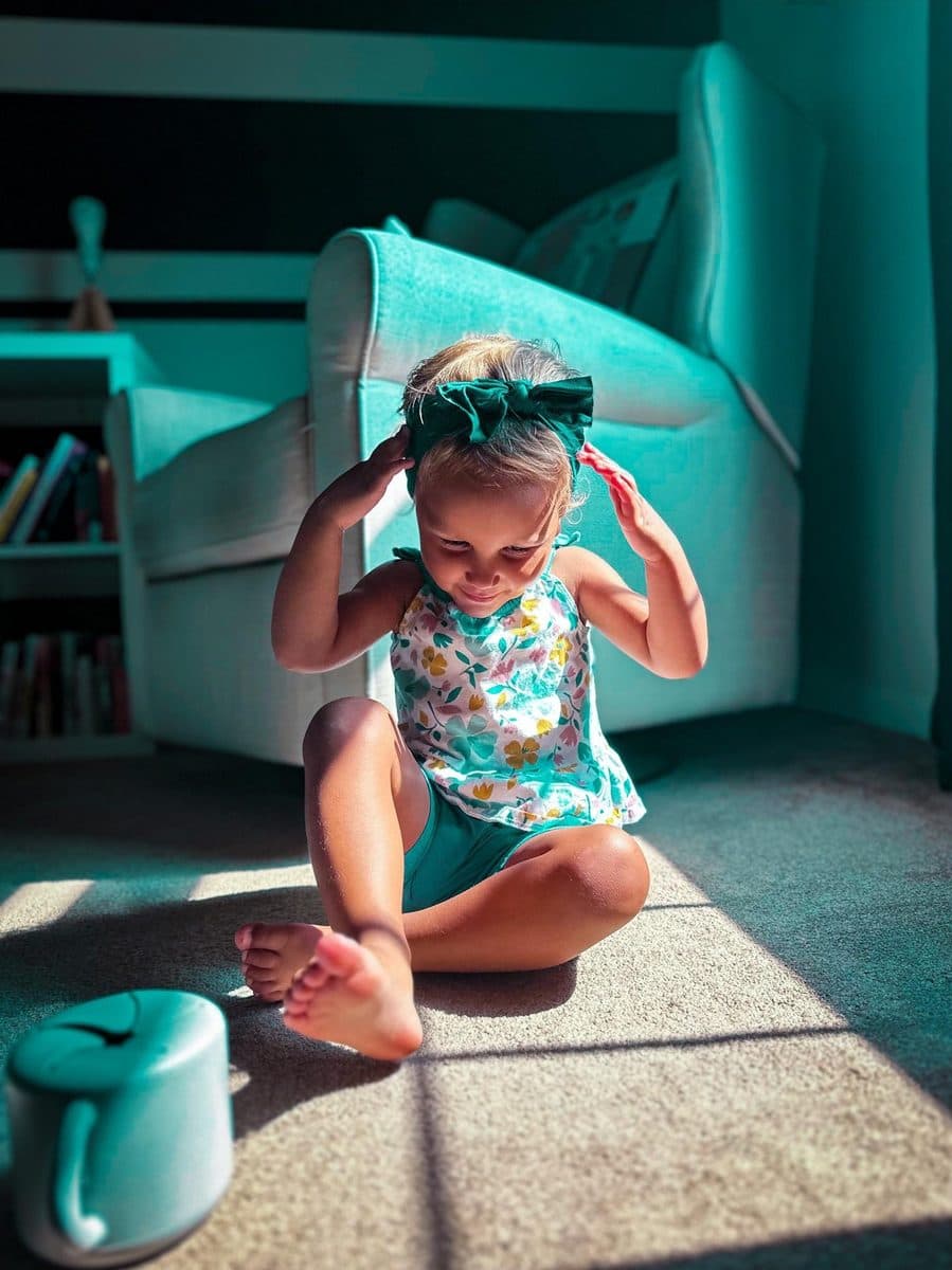 A toddler sits on the carpet in a patch of morning sun, fixing her hair bow