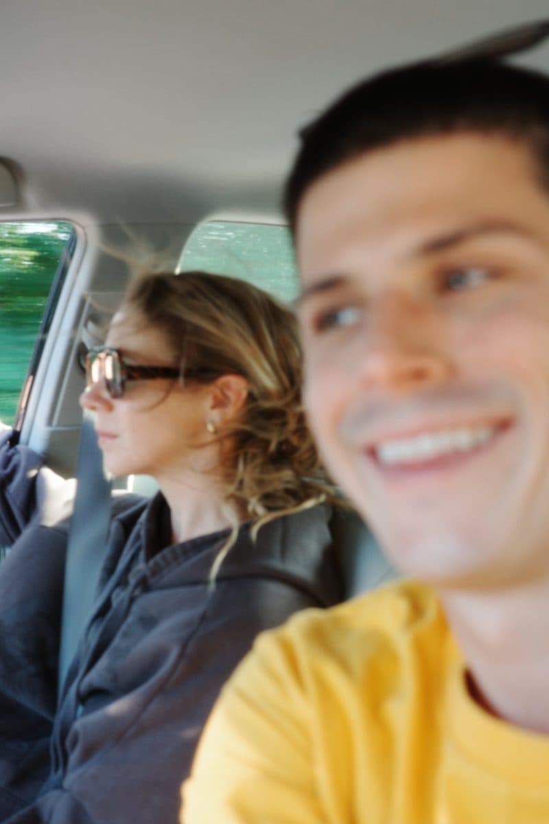 The photographer smiles in the foreground of his own frame in a car