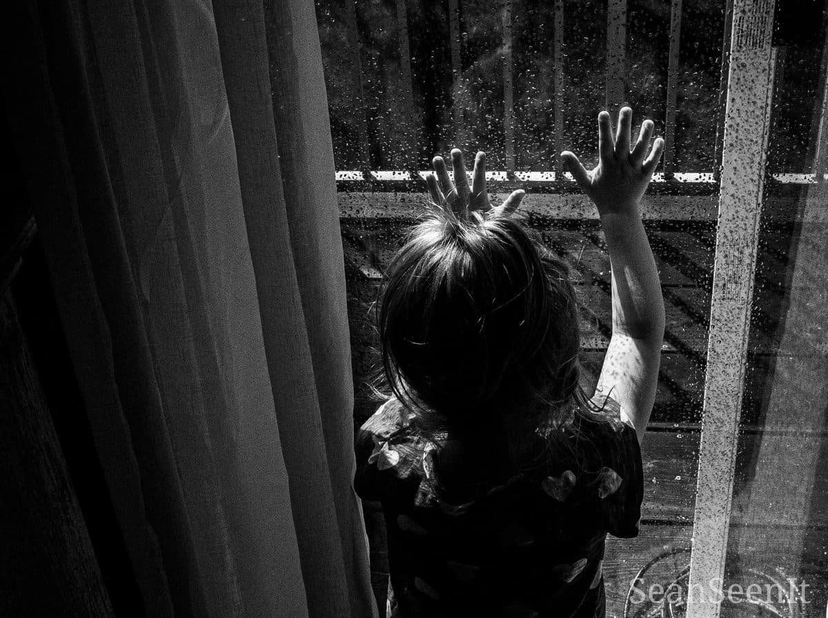 A small girl seen from behind with her hand on a rainy window, black and white
