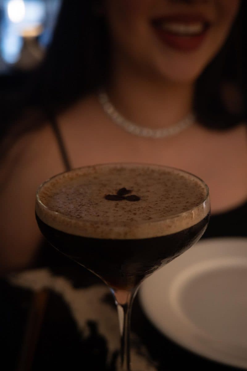 An espresso martini in the foreground with a smiling woman behind it