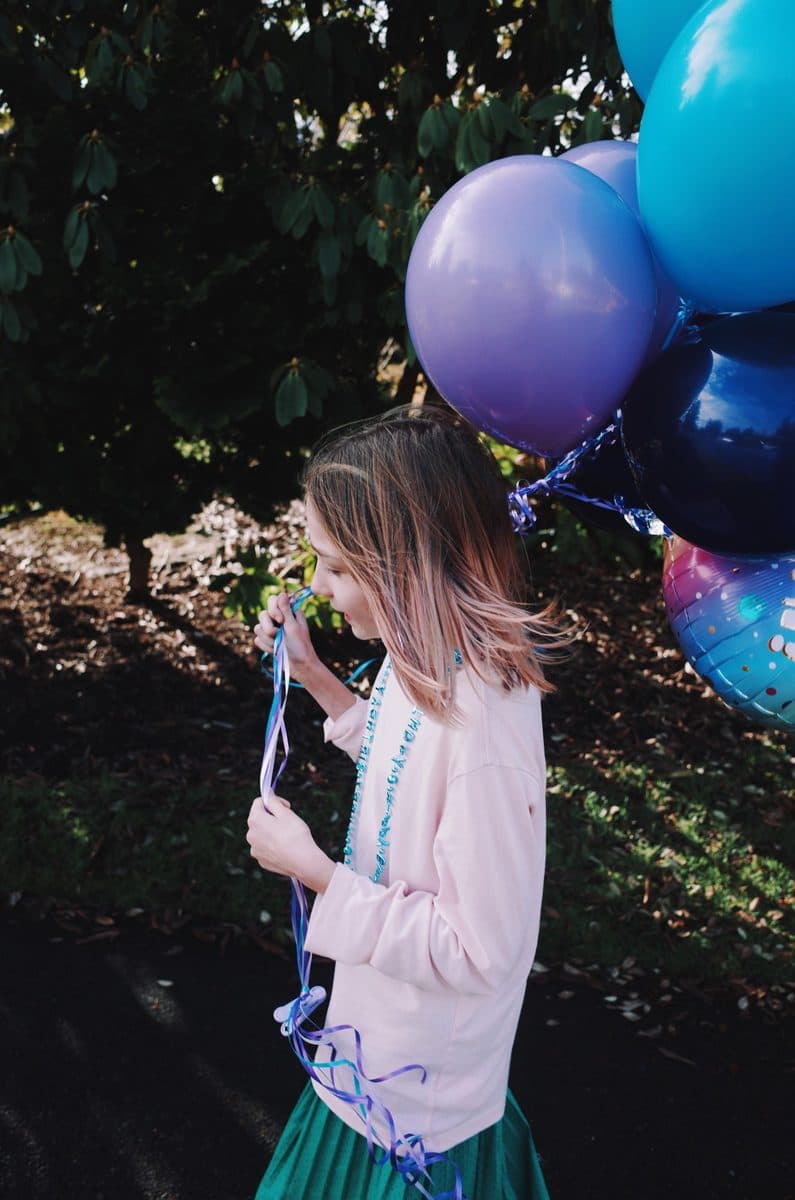 A girl in a pink jacket walks with a bunch of purple and blue balloons