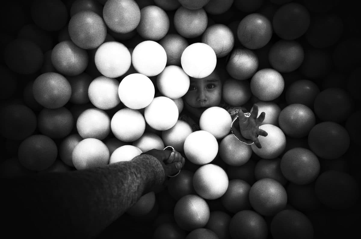 A hand reaches into a ball pit toward a girl peeking out between the balls, black and white