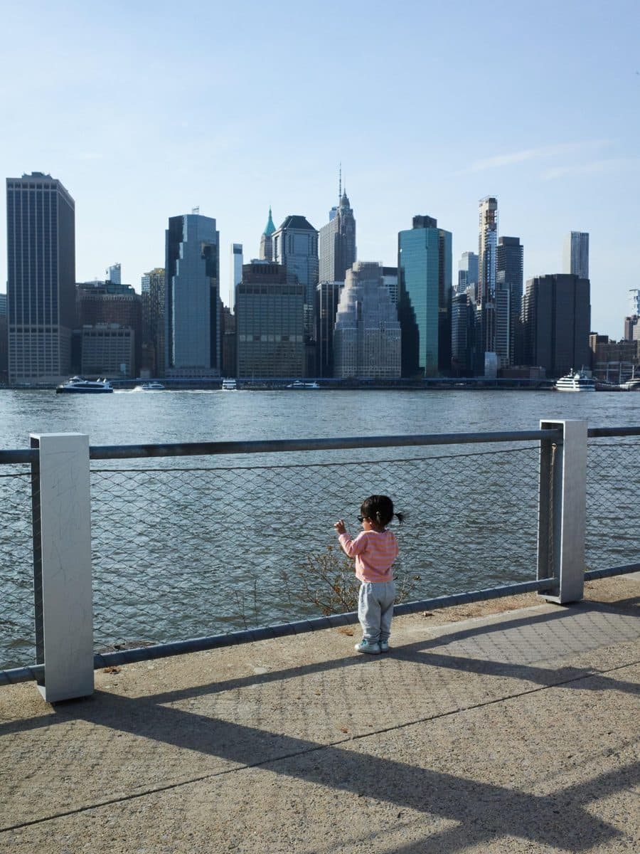 A small girl in pink stands at a river railing with the Manhattan skyline beyond
